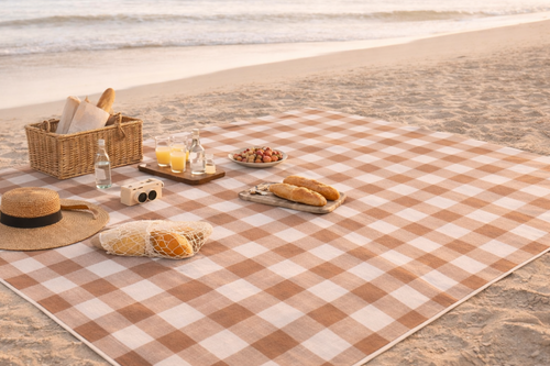 Checkered beach blanket with food and a hat on a sandy beach.